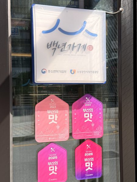 출입문 옆 유리에 걸린 백년가게 현판과 그 아래 붙은 분홍색 ‘부산의 맛’ 스티커 4장. 스티커에는 각각 2023, 2024, 2025, 2026과 부산광역시가 적혀 있다.