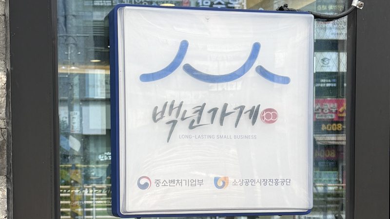 매장 출입문 유리에 걸린 백년가게 현판. 흰 바탕에 ‘백년가게 LONG-LASTING SMALL BUSINESS’ 글자와 중소벤처기업부·소상공인시장진흥공단 표시가 있다.
