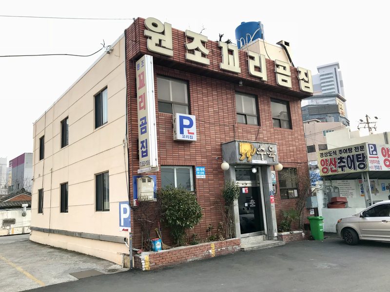 과거 원조꼬리곰집 건물 외관. 붉은 벽돌 2층 건물 옥상에 ‘원조꼬리곰집’ 글자 간판이 있고, 세로 간판과 주차 표시판, 입구 위 ‘수육’ 간판이 보인다.