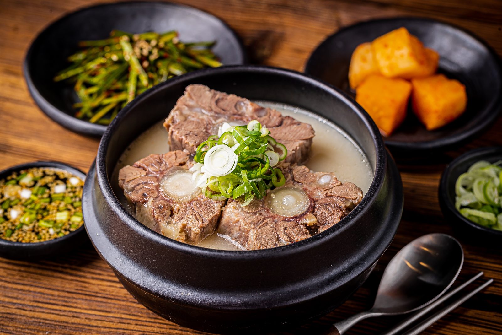 검은 뚝배기에 담긴 꼬리탕. 뽀얀 국물 위에 뼈가 붙은 소꼬리 세 토막과 송송 썬 대파가 올라가 있고, 깍두기·부추무침·양념장이 곁들여져 있다.