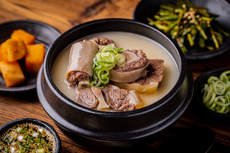 검은 뚝배기에 담긴 한우꼬리탕. 뽀얀 국물에 꼬리와 함께 여러 부위의 고기가 들어 있고 대파가 올라가 있다. 옆에 깍두기와 부추무침이 있다.