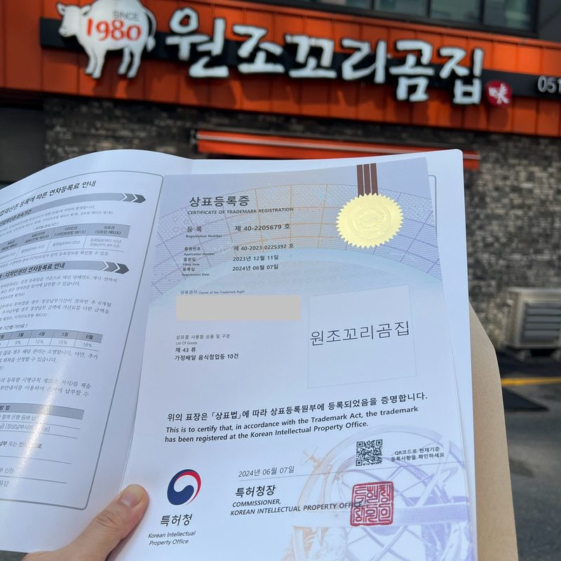 원조꼬리곰집 간판 앞에서 손에 든 특허청 상표등록증. 등록번호 제40-2205679호, 출원번호 제40-2023-0225392호, 출원일 2023년 12월 11일, 등록일 2024년 6월 7일, 상표 ‘원조꼬리곰집’, 제43류가 적혀 있고 상표권자 칸은 가려져 있다.